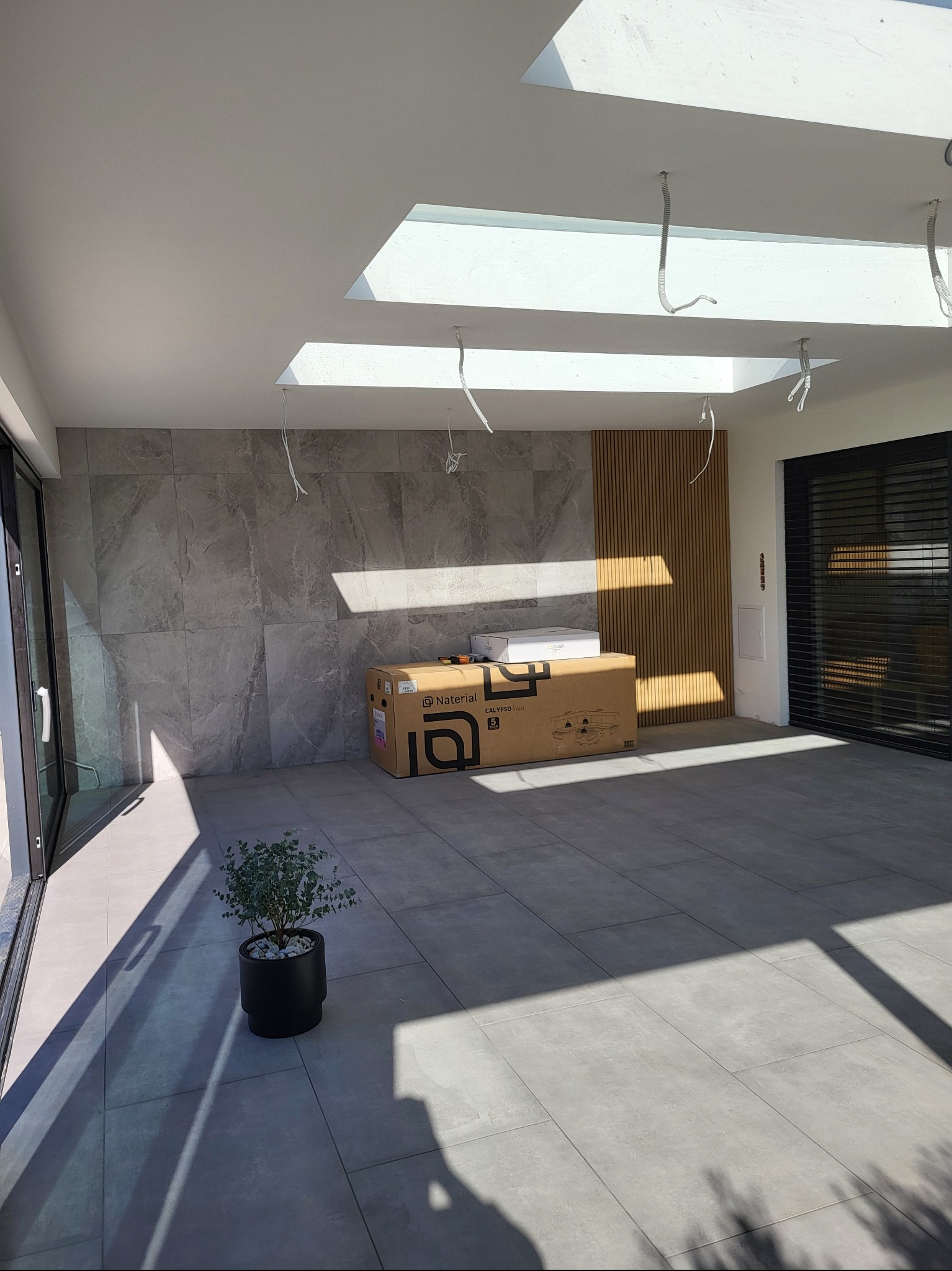 Jasne wnętrze z szarymi płytkami podłogowymi i ściennymi, drewnianą ścianką działową, otwartym oknem i rośliną w doniczce. W tle kartonowe pudło.