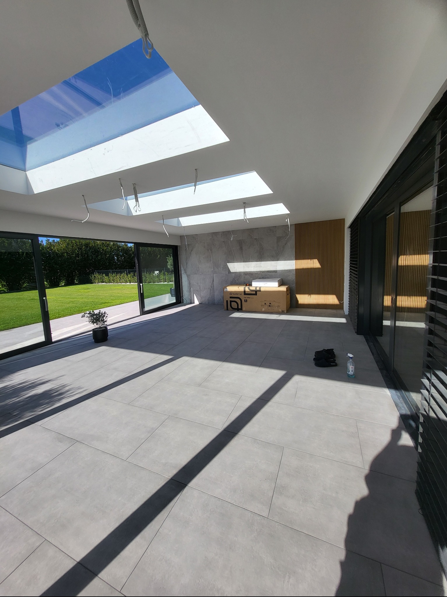 Jasne wnętrze z dużymi oknami i szarymi płytkami podłogowymi. Widoczne elementy dekoracyjne z drewna i karton z napisem 'Material'.