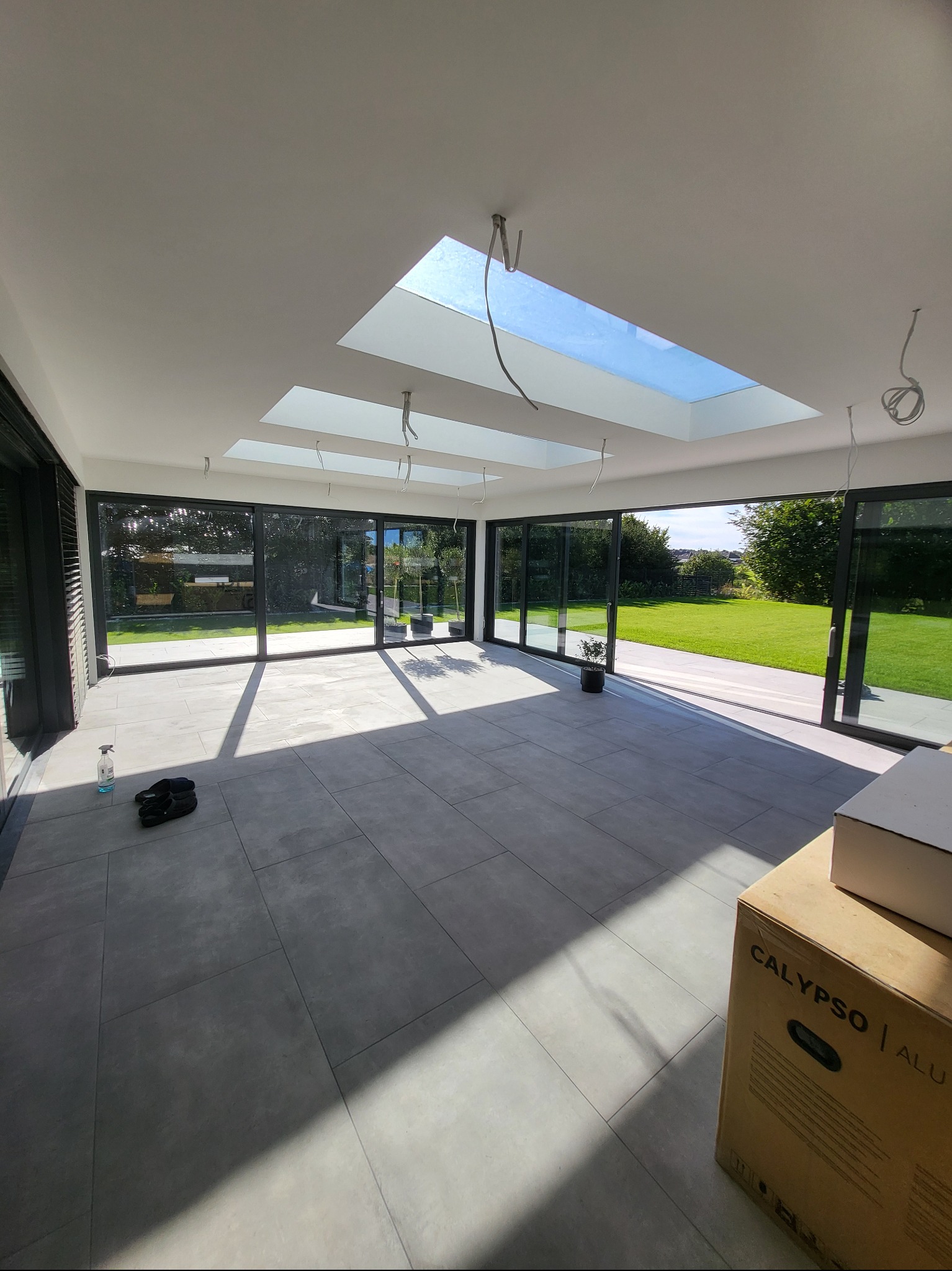 Jasne wnętrze z dużymi oknami przesuwnymi i szarymi płytkami podłogowymi. Widoczne trzy okna dachowe i elementy instalacji elektrycznej. Na podłodze leżą buty i stoi karton.