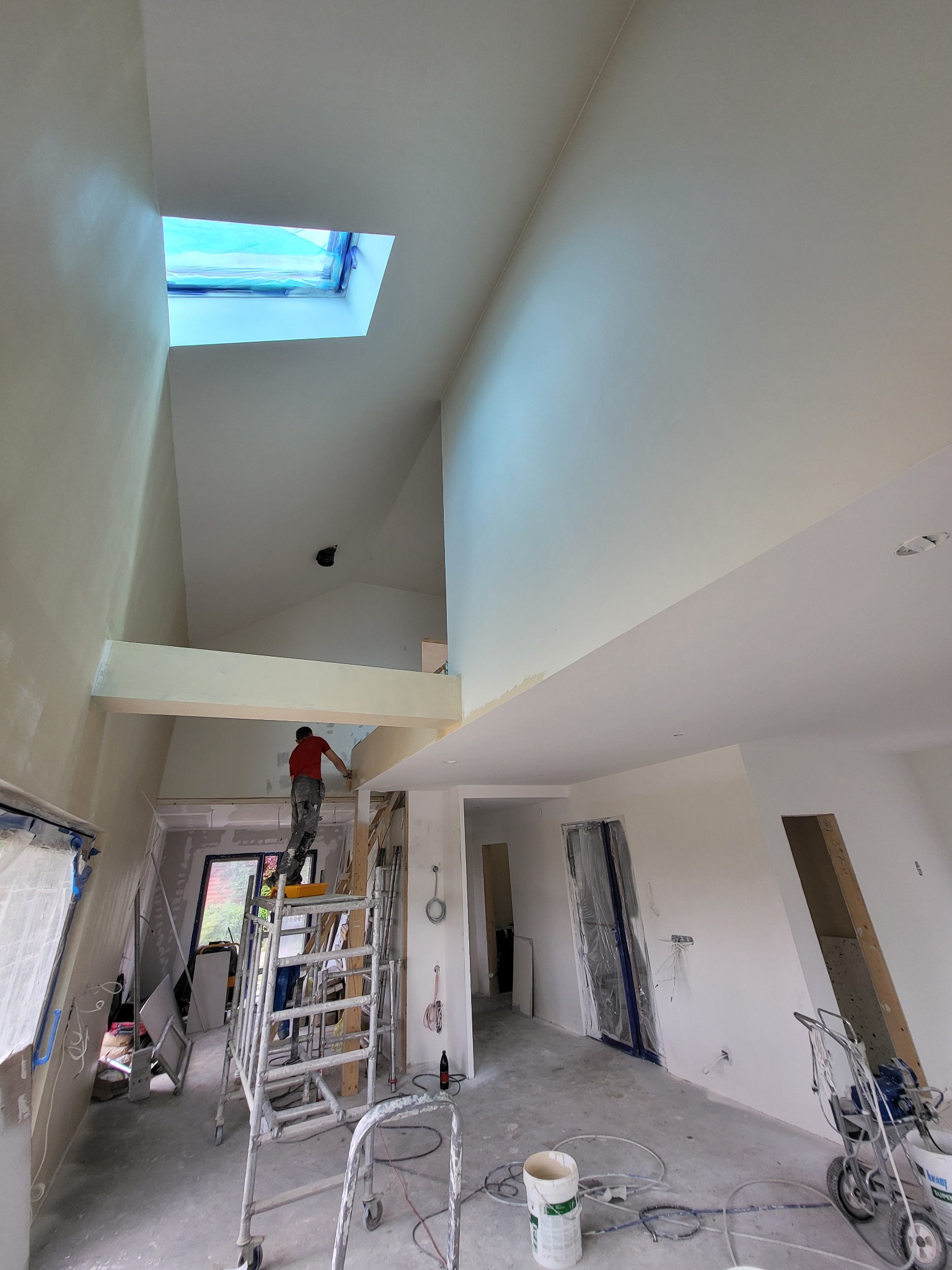 Wysoki pokój ze skośnym dachem w trakcie malowania. Malarz na rusztowaniu maluje ścianę pod oknem dachowym. Widoczne narzędzia malarskie i zabezpieczone elementy.