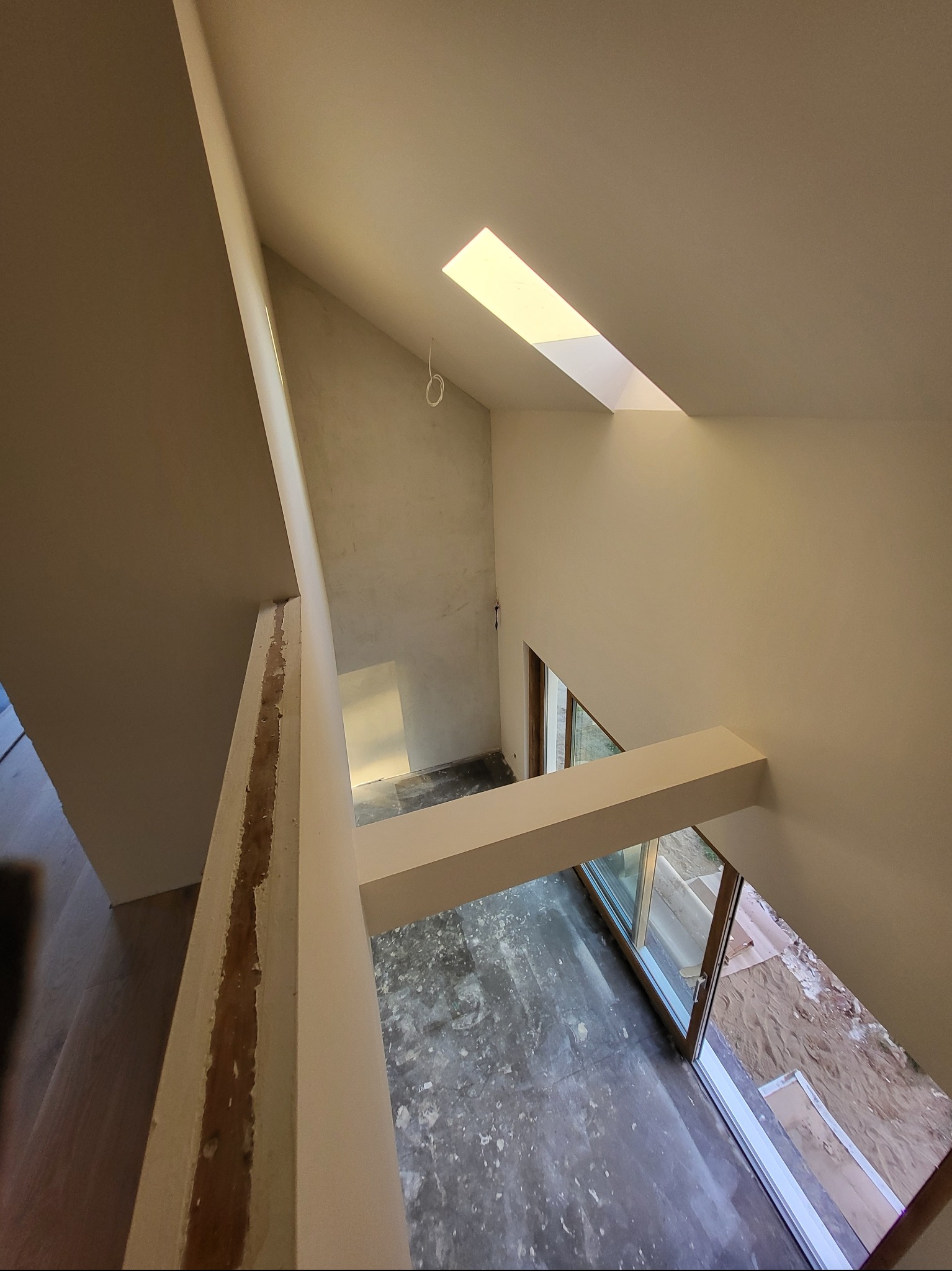 Wnętrze domu w trakcie prac wykończeniowych: surowe ściany, okno dachowe wpuszczające światło, widok z góry na otwarte okno balkonowe i podłogę przygotowaną do dalszych prac.