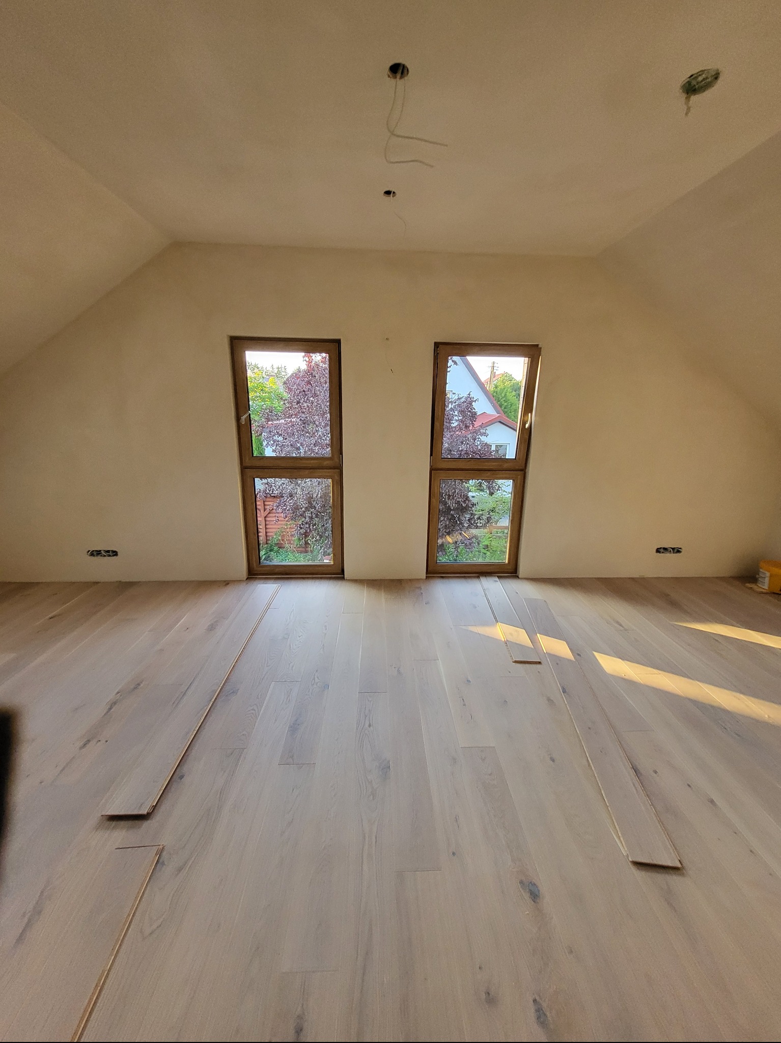 Montaż jasnej, drewnianej podłogi w pokoju na poddaszu z dwoma oknami. Widoczne deski podłogowe w trakcie układania, surowe ściany i przygotowane punkty elektryczne.