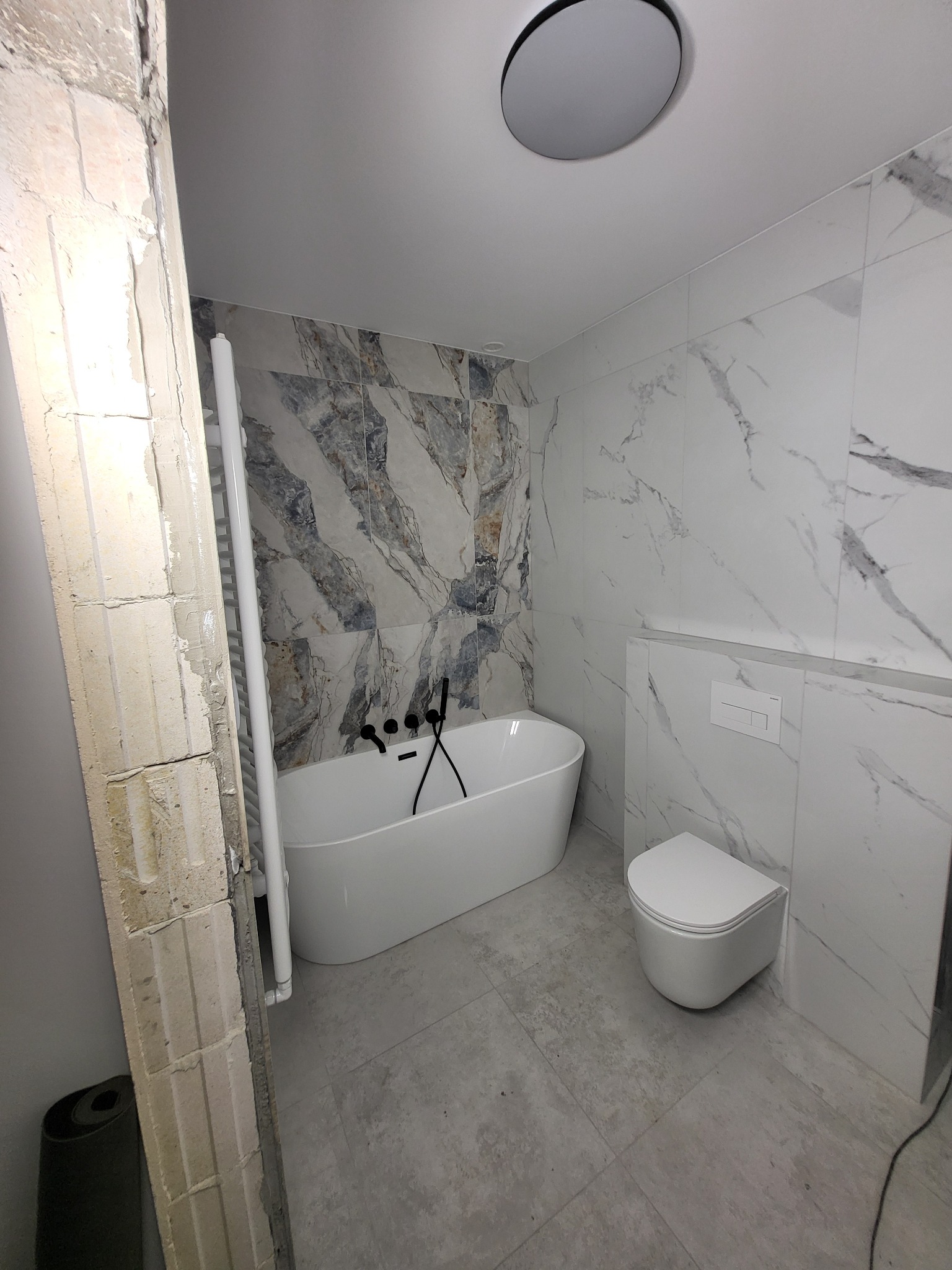 Nowoczesna łazienka w trakcie remontu: biała wanna wolnostojąca, podwieszana toaleta, marmurowe płytki na ścianach, surowy filar z cegieł. Widoczne etapy prac wykończeniowych.