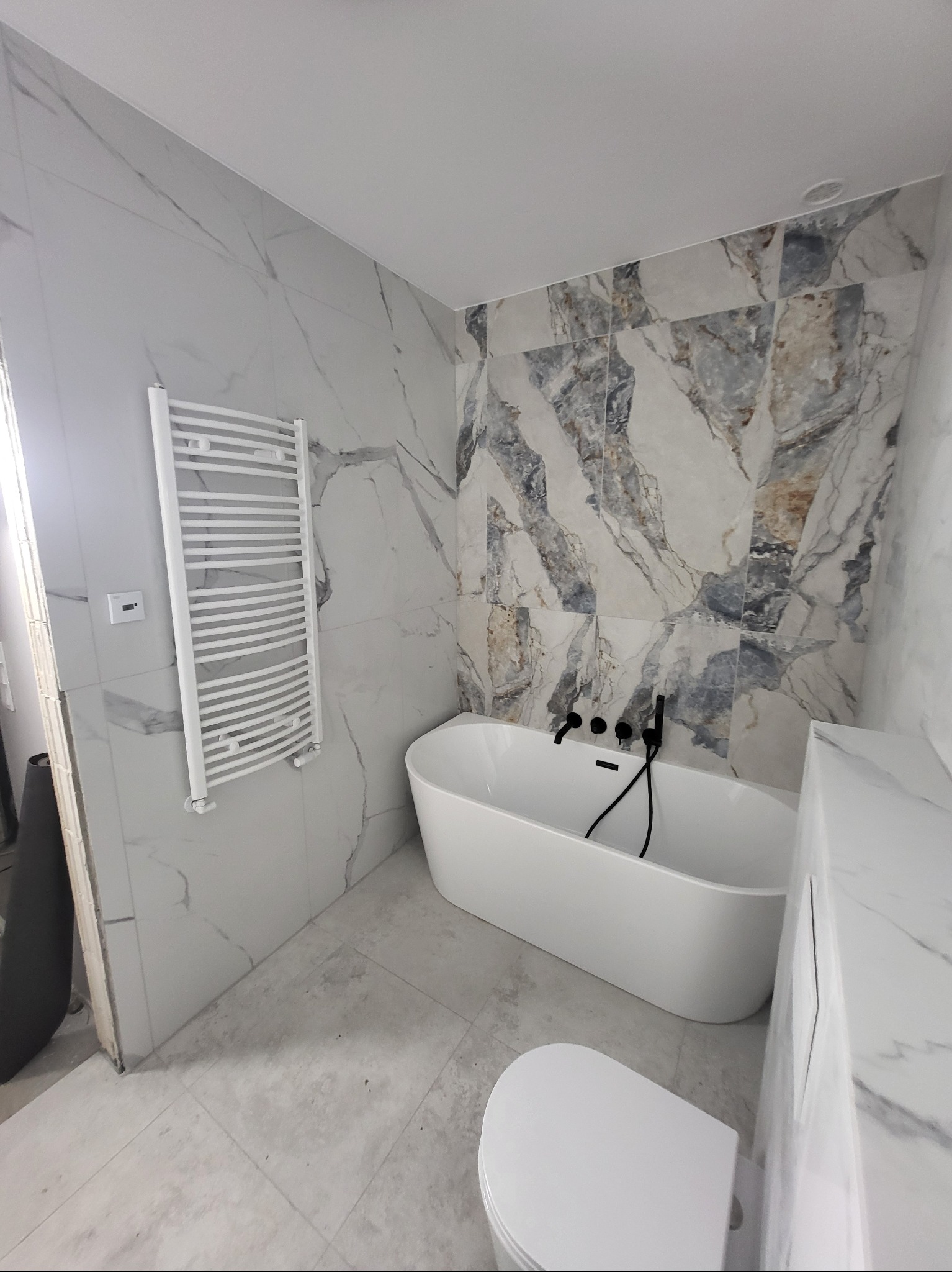 Wykończona łazienka z wanną wolnostojącą, grzejnikiem drabinkowym i marmurowymi płytkami w odcieniach bieli i szarości. Czarna armatura dodaje elegancji.