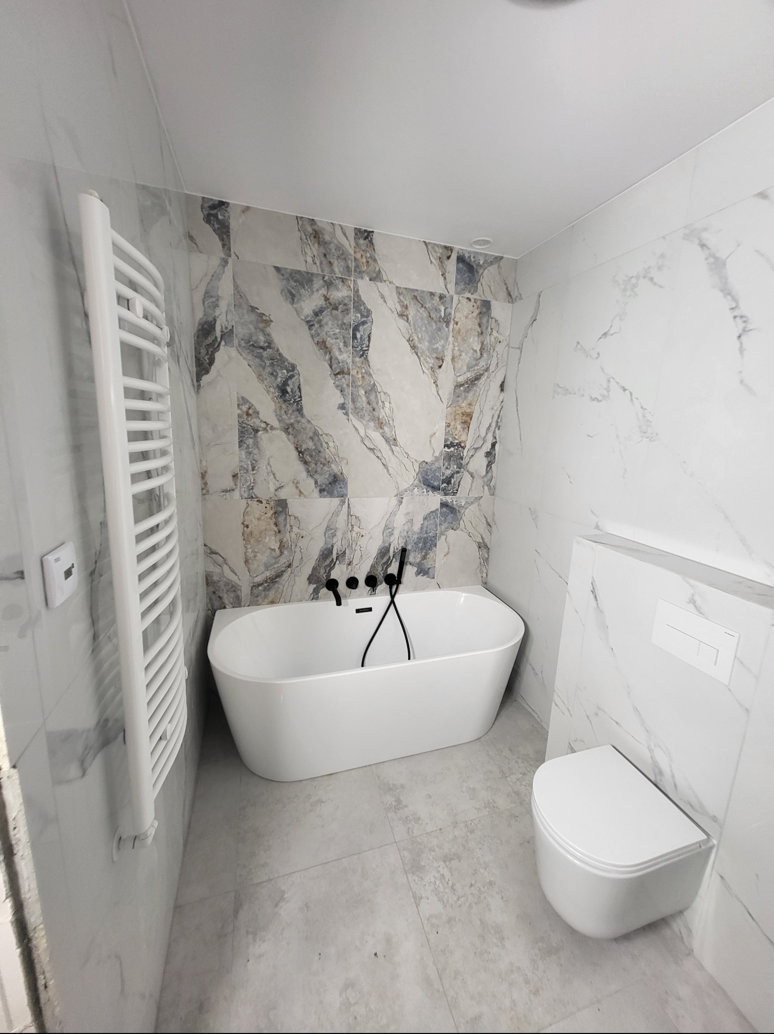 Nowoczesna łazienka z wolnostojącą wanną, grzejnikiem drabinkowym i podwieszaną toaletą. Marmurowe płytki na ścianach i podłodze. Czarna armatura.