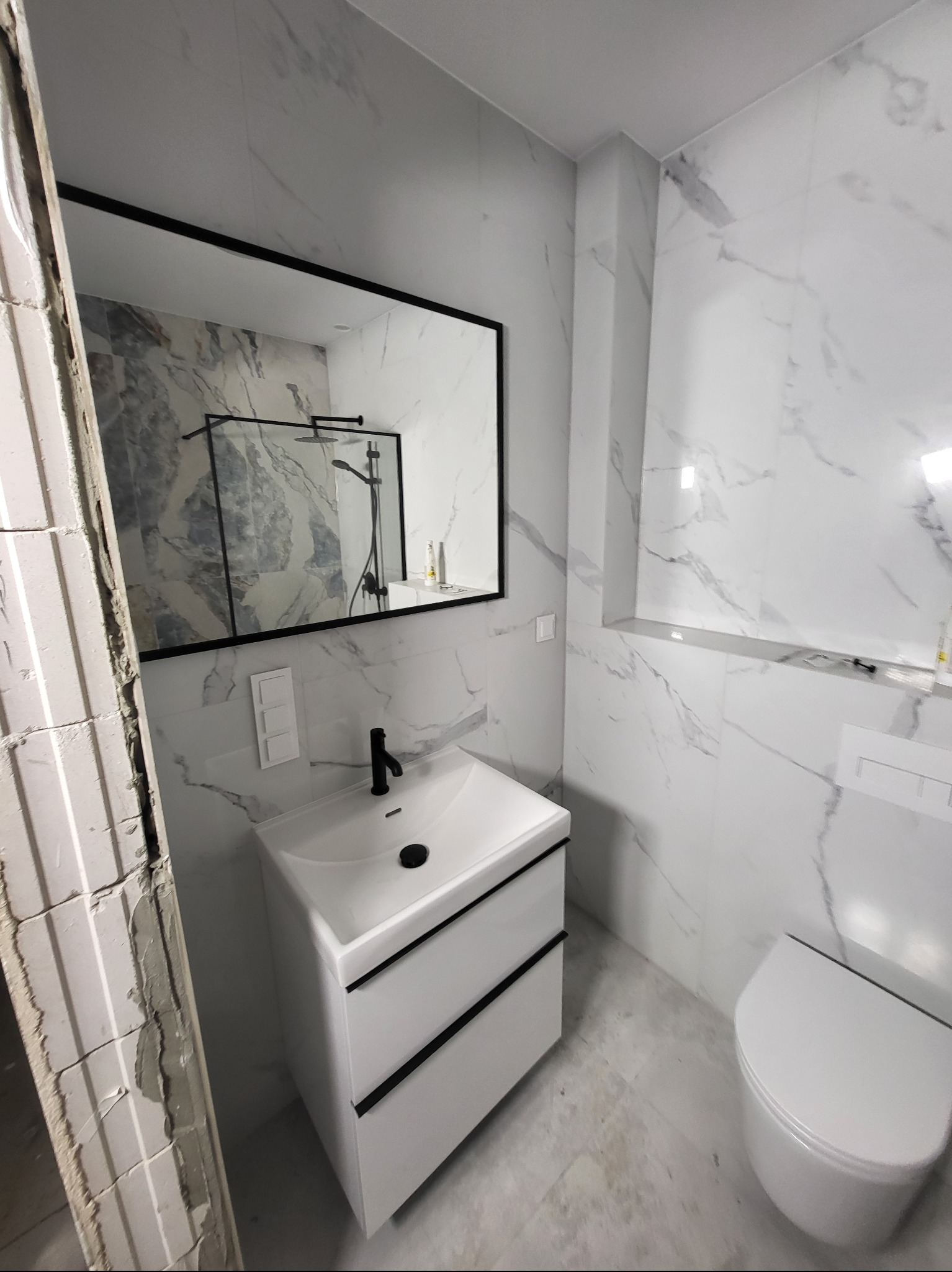 Nowoczesna łazienka w trakcie wykończenia: białe marmurowe płytki, czarna armatura, wisząca toaleta i fragment ściany w stanie surowym.