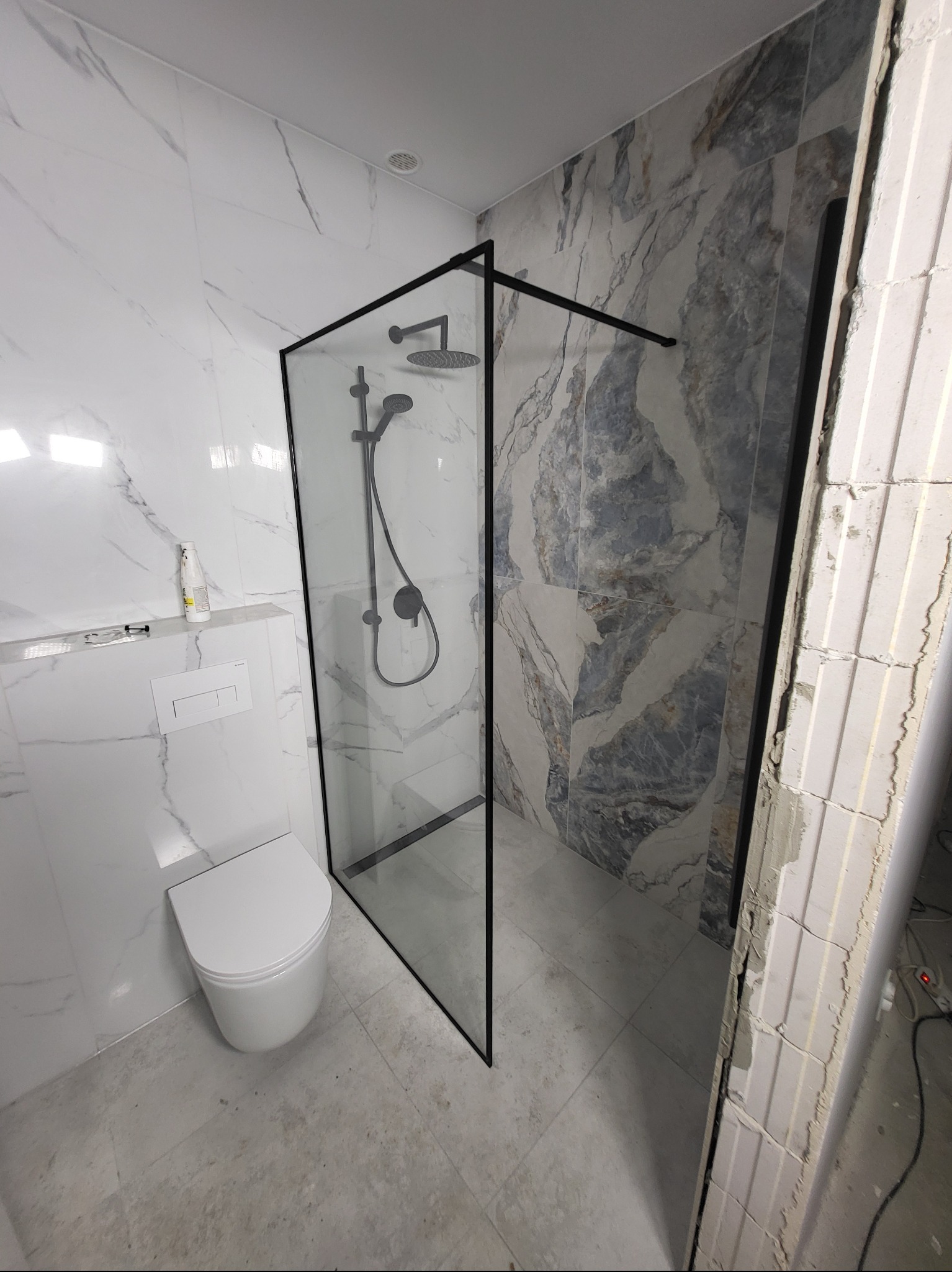 Nowoczesna łazienka z kabiną prysznicową typu walk-in z czarnymi profilami, marmurowe płytki na ścianach i podłodze, wisząca toaleta. Częściowo widoczne niedokończone prace.