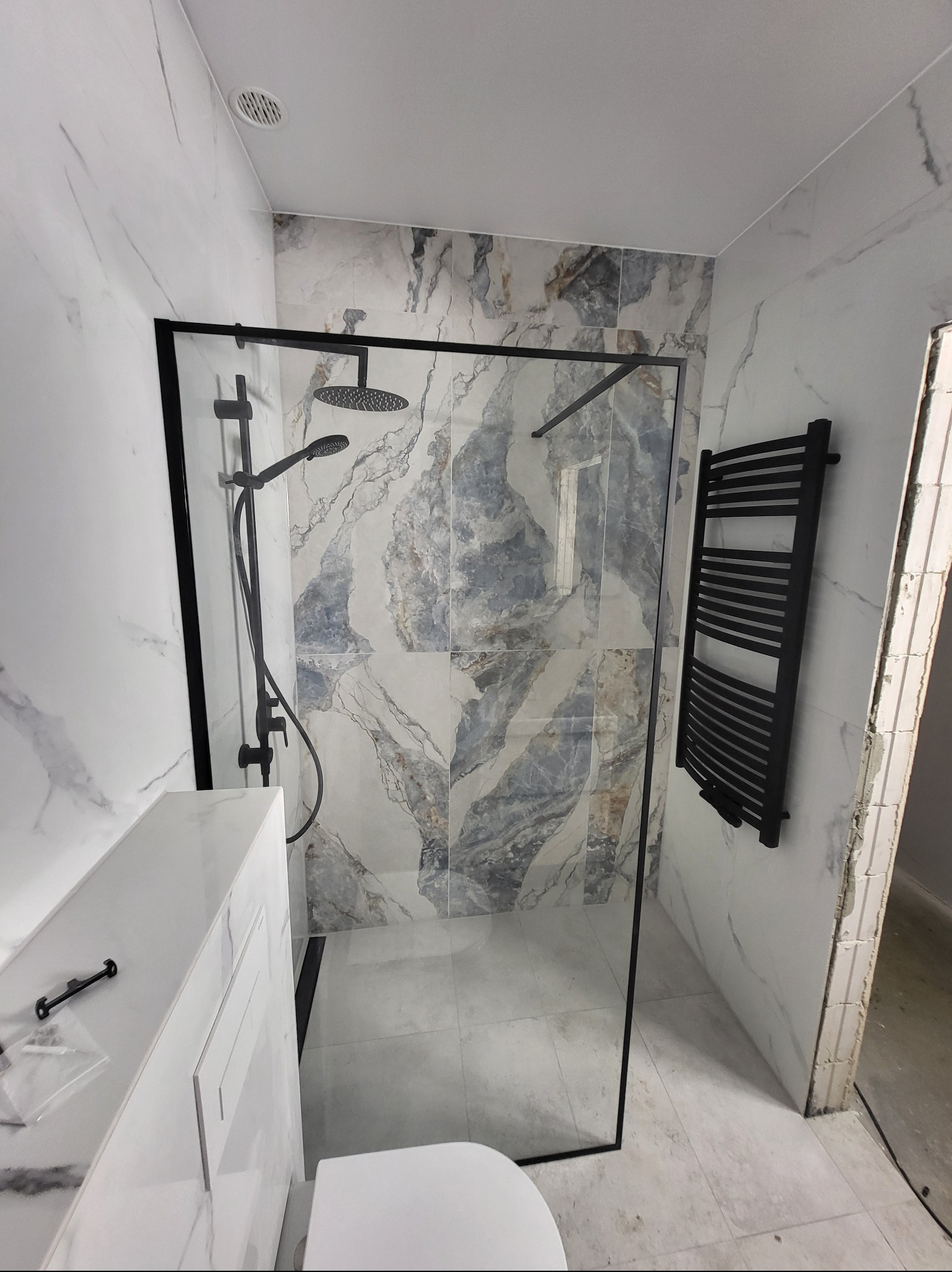 Nowoczesna łazienka z kabiną prysznicową typu walk-in, marmurowe płytki na ścianach i podłodze, czarna armatura i grzejnik drabinkowy. Widok na fragment toalety.