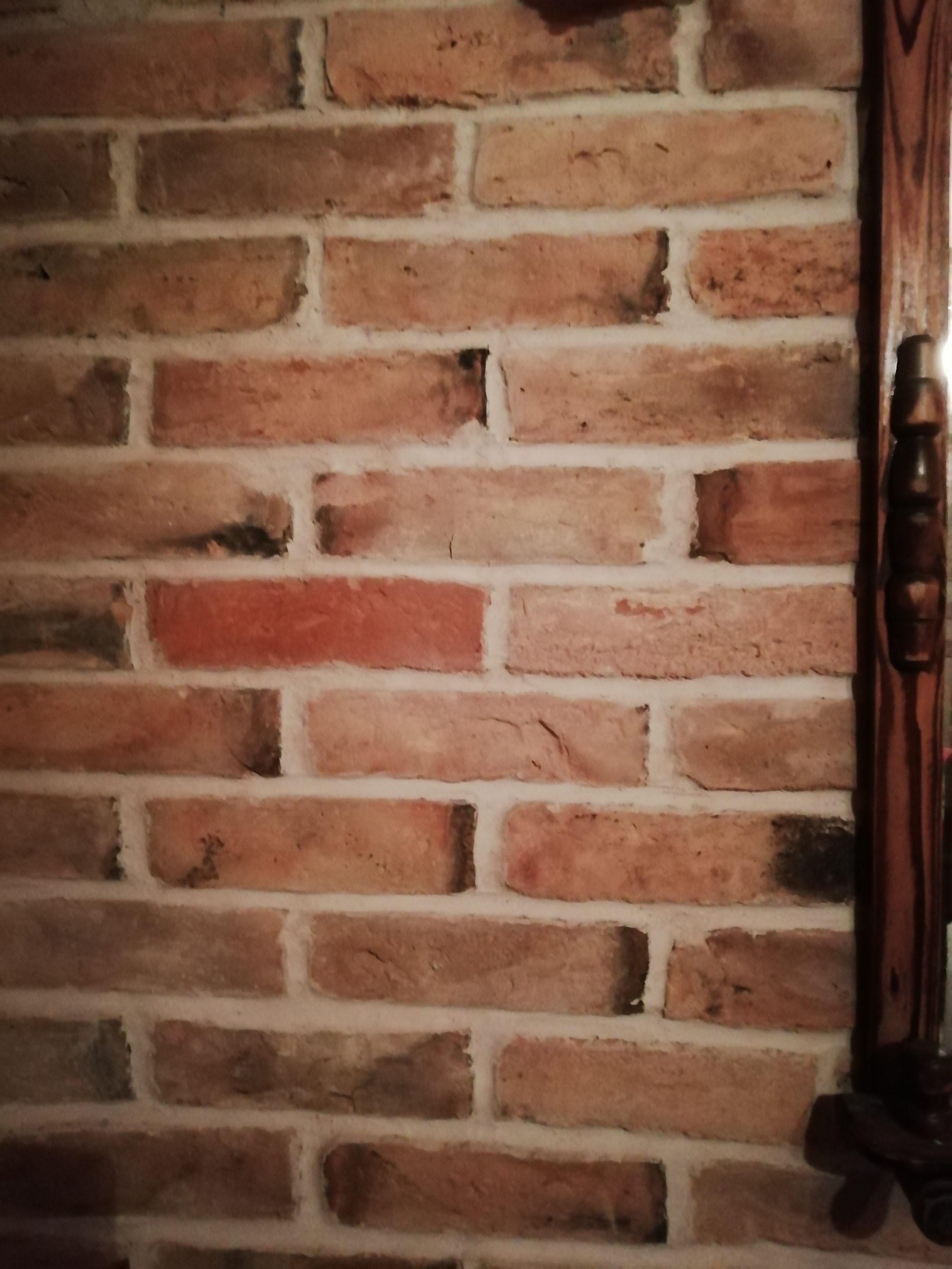 Fragment ceglanej ściany w odcieniach czerwieni i brązu, z widoczną drewnianą kolumną po prawej stronie, tworzącą element dekoracyjny.