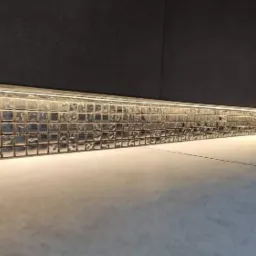 Rząd błyszczących, kwadratowych płytek mozaikowych z oświetleniem LED, ułożonych wzdłuż ściany nad podłogą z szarych płytek, tworzących dekoracyjny akcent w pomieszczeniu.