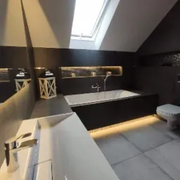 Nowoczesna łazienka na poddaszu z czarnymi płytkami, białą wanną i umywalką, oświetleniem LED pod wanną i w niszy ściennej, oraz dekoracyjną latarnią odbijającą się w lustrze.