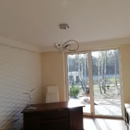 Nowoczesne biuro z dekoracyjną ścianą 3D, ciemnym biurkiem i widokiem na ogród przez duże okno.