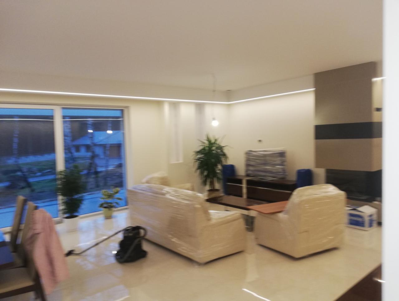 Salon po remoncie z meblami zabezpieczonymi folią, widoczne oświetlenie LED podsufitowe, rośliny doniczkowe przy oknie i odkurzacz na błyszczącej podłodze.