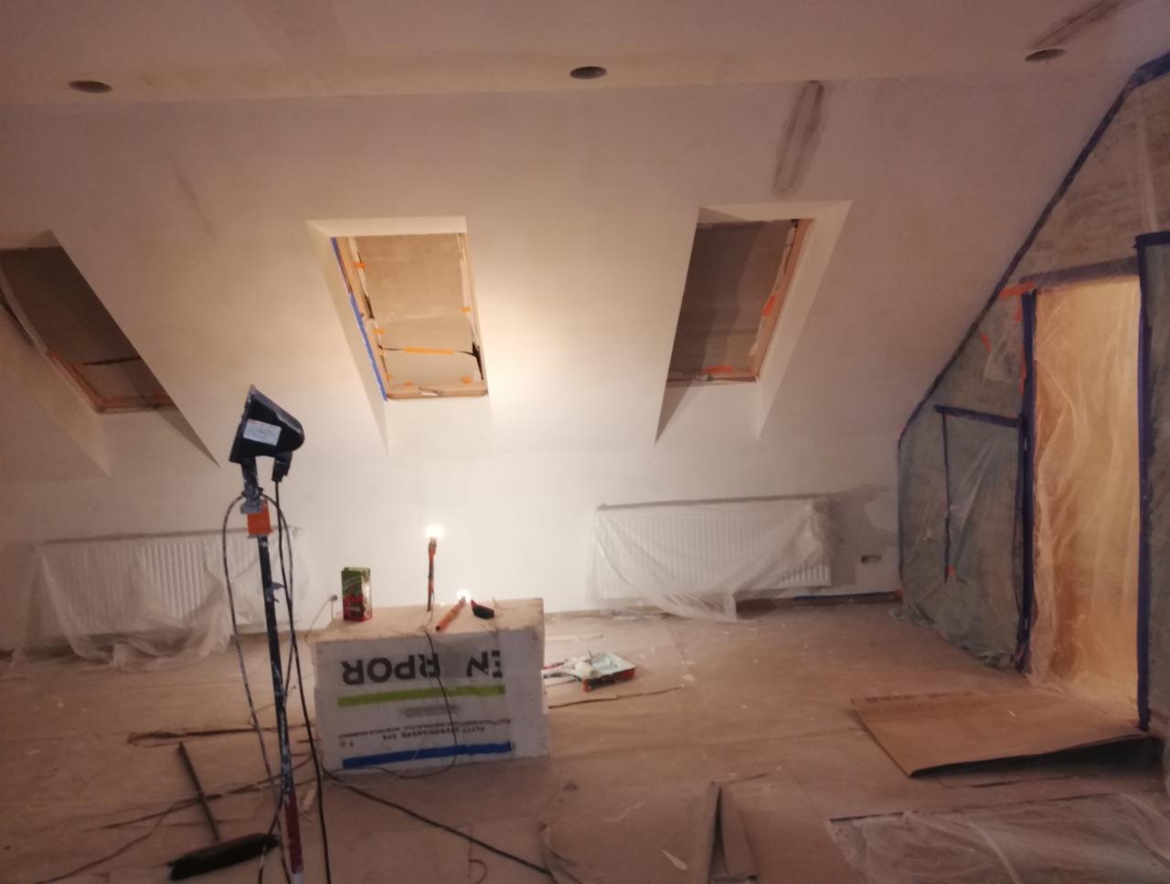 Pokój na poddaszu w trakcie prac wykończeniowych, widoczne okna dachowe z taśmą malarską, zabezpieczone grzejniki i drzwi folią, lampa halogenowa na statywie i styropianowy blok.