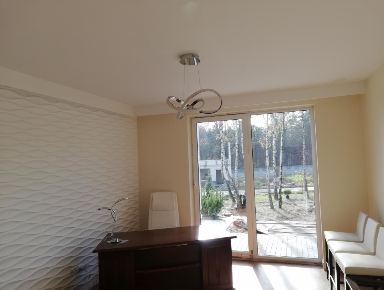 Nowoczesne biuro z dekoracyjną ścianą 3D, ciemnym biurkiem i widokiem na ogród przez duże okno.