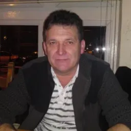 Portret mężczyzny w swetrze i koszulce w paski, siedzącego przy stole z pustą szklanką i zegarkiem na ręku, na tle okna z żaluzjami.