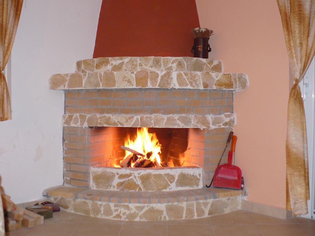 Tradycyjny kominek z cegły i kamienia w salonie, z płomieniami ognia, pogrzebacz i czerwona szufelka obok, dekoracyjny element na górze.