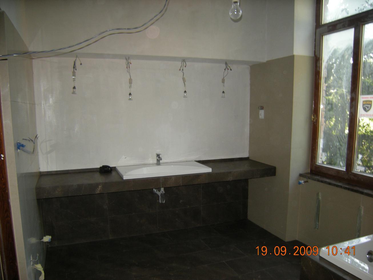 Łazienka w trakcie remontu: widoczne płytki na podłodze i obudowie umywalki, surowe ściany z wystającymi kablami, niedokończone oświetlenie i okno z drewnianą ramą.