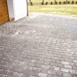 Ułożona szara kostka brukowa na podjeździe, widok z poziomu gruntu, brama garażowa z lewej strony, trawnik w tle.