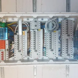 Otwarta skrzynka rozdzielcza z zainstalowanymi bezpiecznikami, przewodami i szynami DIN, widoczne elementy zabezpieczeń przepięciowych i r&oacute;żnicowoprądowych, kable ułożone w pętlę.