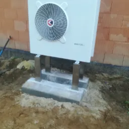 Biała jednostka zewnętrzna z logo Chaffoteaux, zamontowana na metalowych wspornikach na betonowej podstawie, widoczna na tle ceglanej ściany w trakcie instalacji.