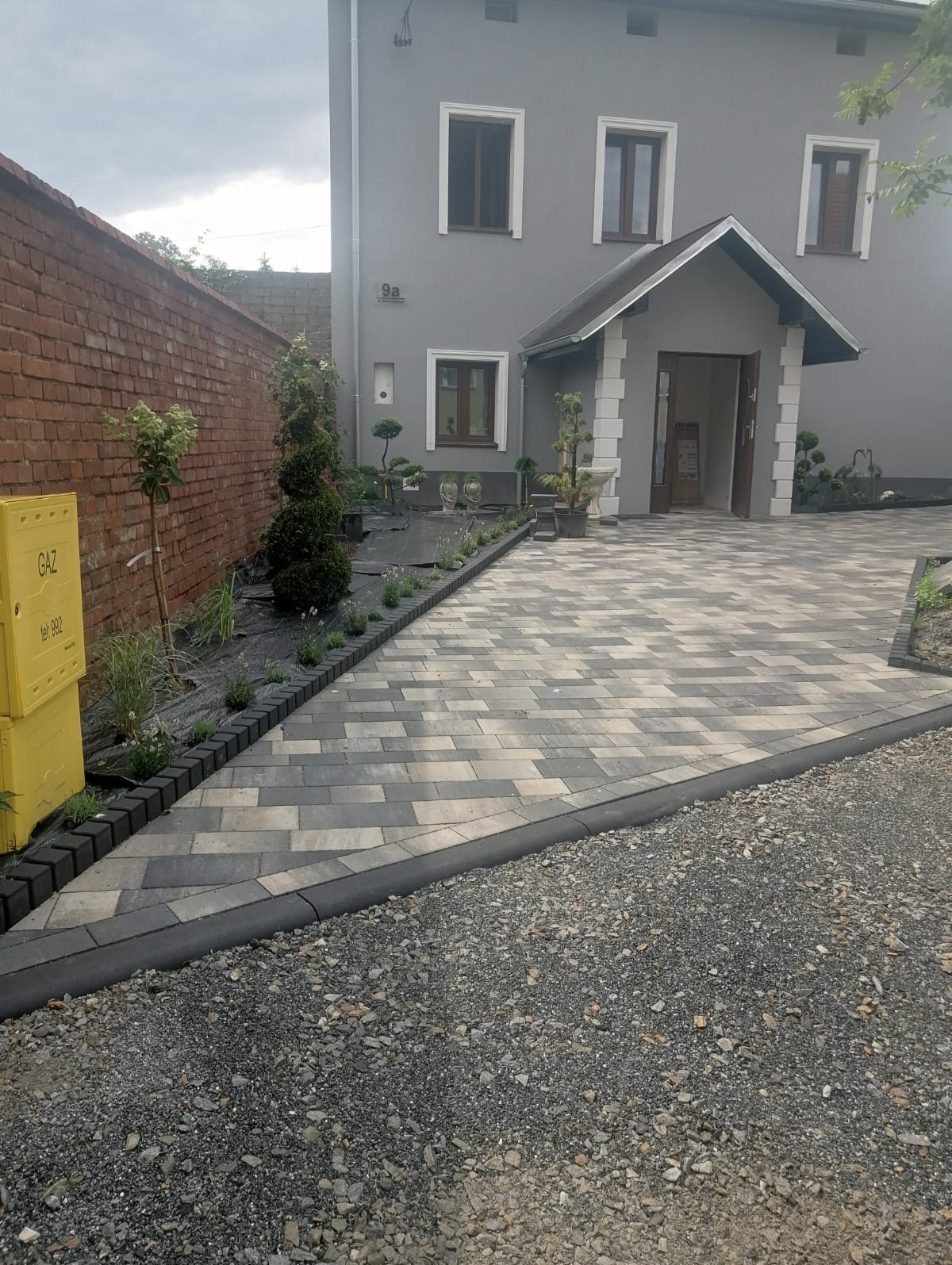 Ułożona kostka brukowa w odcieniach szarości i beżu przed domem z szarą elewacją, widoczny żółty skrzynka gazowa i obrzeża z kostki przy rabatach z roślinami.