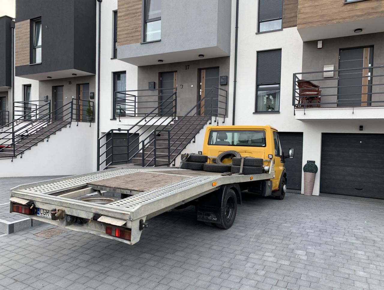 Żółta laweta z kilkoma oponami na platformie, zaparkowana na brukowanym podjeździe przed nowoczesnymi domami szeregowymi z numerami 15 i 17.