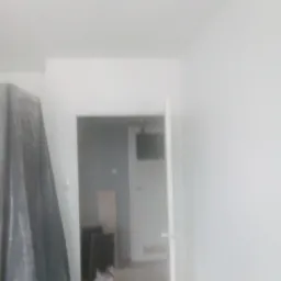 Przygotowanie do prac w Grodzisku Mazowieckim: Pokryta folią meblową część pokoju, widok na odświeżone, białe ściany i otwarte drzwi do kolejnego pomieszczenia.