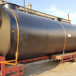 Czarny, cylindryczny zbiornik przemysłowy z włazem, zabezpieczony pomarańczowymi pasami transportowymi na czerwonej platformie niskopodwoziowej, na tle hali przemysłowej.