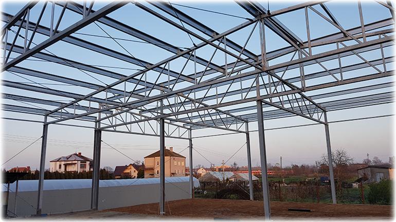 Konstrukcja stalowa hali w trakcie budowy, widoczne elementy nośne i poprzeczne, w tle zabudowa jednorodzinna i zachodzące słońce.
