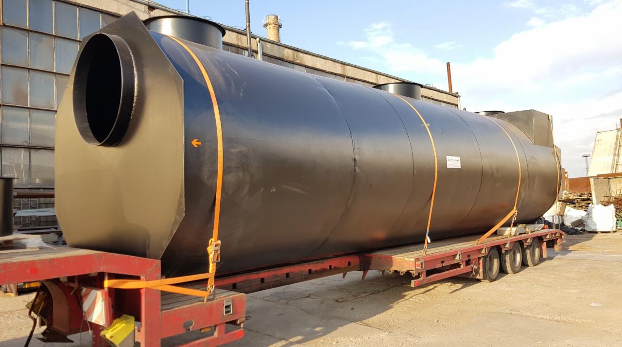 Czarny, cylindryczny zbiornik przemysłowy z włazem, zabezpieczony pomarańczowymi pasami transportowymi na czerwonej platformie niskopodwoziowej, na tle hali przemysłowej.