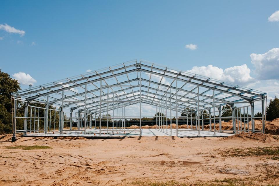 Szkielet hali produkcyjnej z jasnej stali, widoczny na tle błękitnego nieba z białymi chmurami, świeżo wylana betonowa podłoga i ziemia wokół.