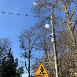 Kamera monitoringu przymocowana do betonowego słupa energetycznego na tle błękitnego nieba i drzew bez liści, obok znak przejścia dla pieszych.