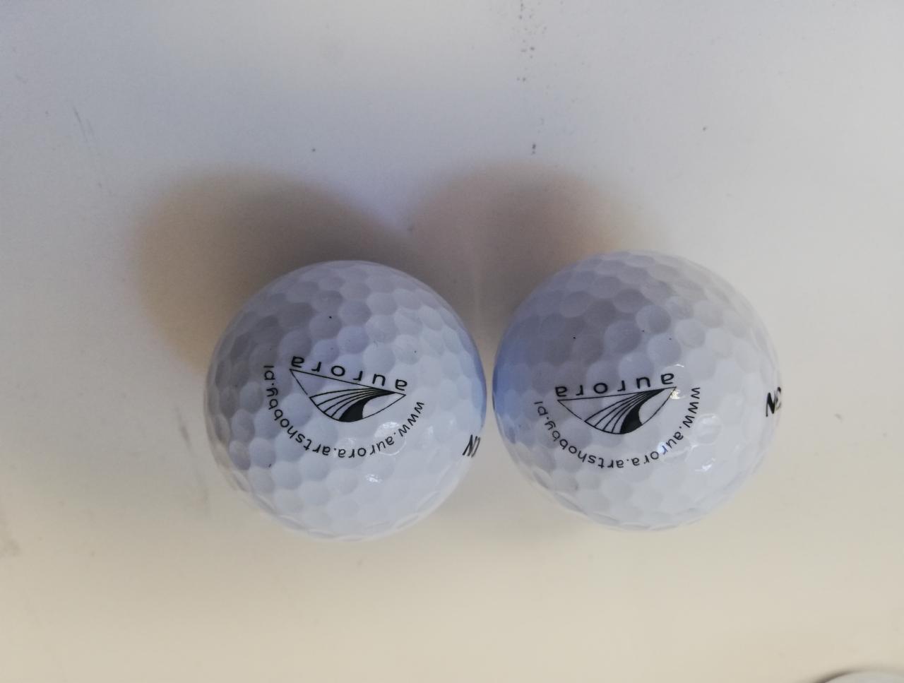 Dwie białe piłeczki golfowe z nadrukowanym logo Aurora i adresem strony internetowej artshobby.pl, leżące na jasnym tle.