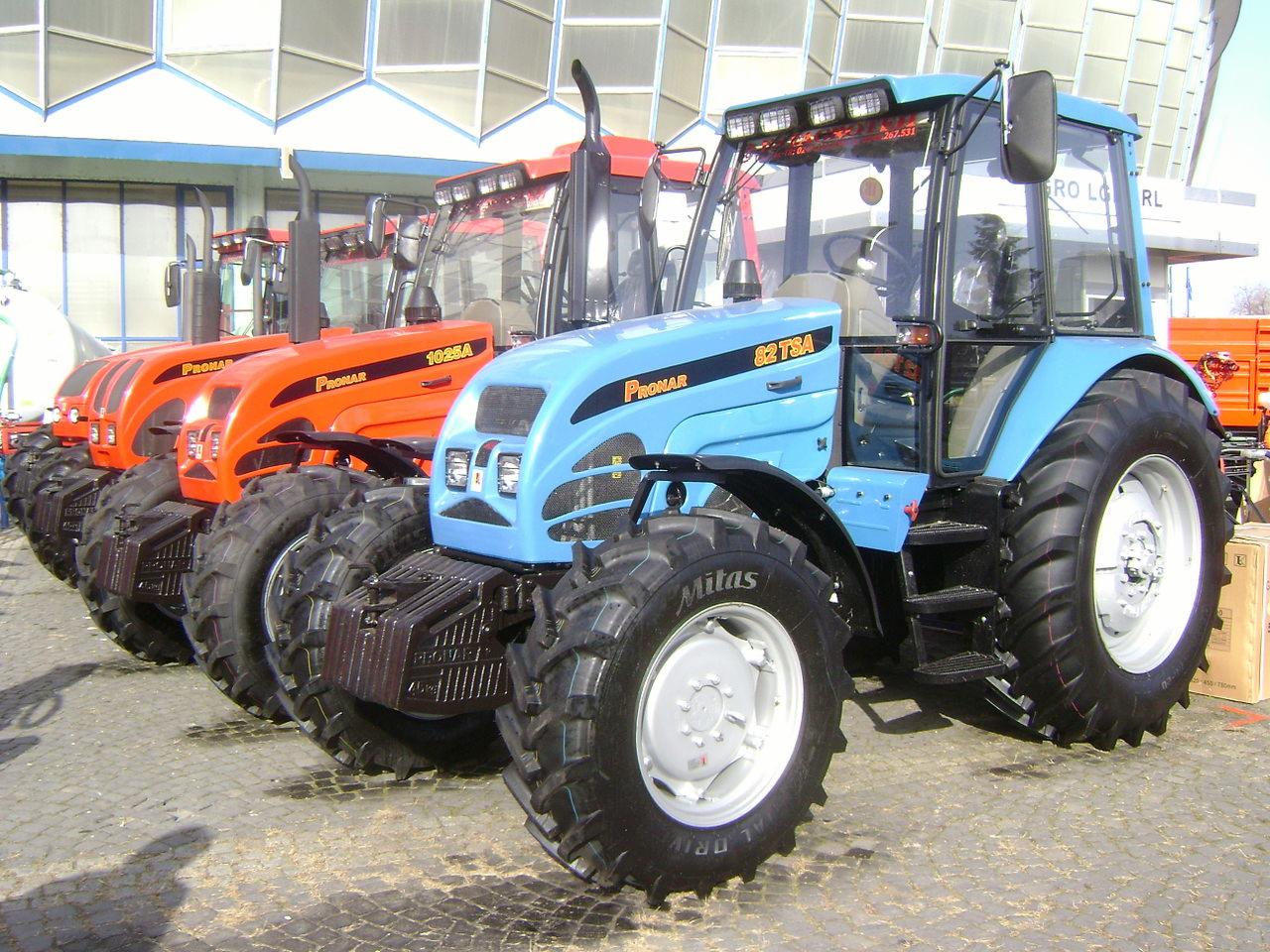 Rząd nowych, pomarańczowych traktorów Pronar 1025A i niebieski Pronar 82 TSA, ustawionych na brukowanym placu przed budynkiem o szklanej fasadzie.