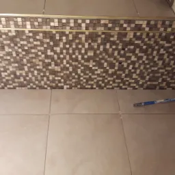 Układanie beżowych płytek na ścianie i podłodze z użyciem krzyżyków dystansowych, widoczny fragment dekoracyjnej mozaiki z brązowymi kwadratami i listwą wykończeniową.