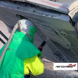 Pracownik w kombinezonie ochronnym i hełmie piaskuje podwozie samochodu, usuwając rdzę i przygotowując do lakierowania. Widoczny efekt czyszczenia i logo firmy Renowacje24.