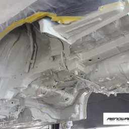 Podwozie samochodu po zabezpieczeniu antykorozyjnym, widoczne świeżo nałożone białe zabezpieczenie, z elementami zakrytymi taśmą malarską i folią ochronną, logo firmy w prawym dolnym rogu.