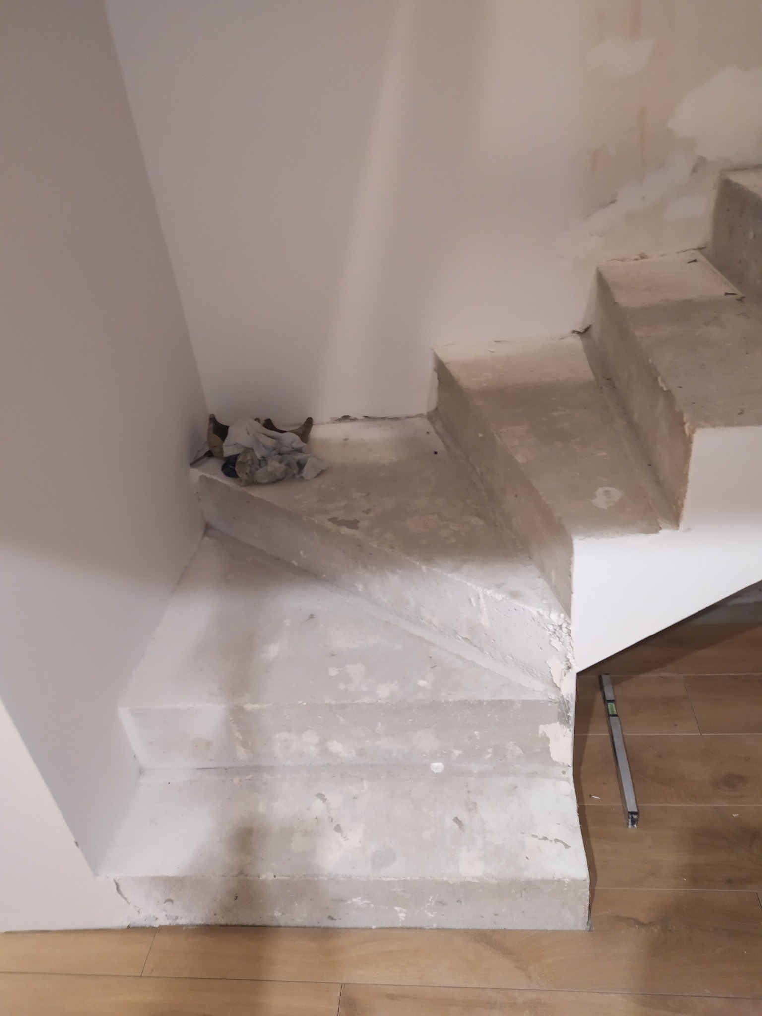 Surowe betonowe schody wewnętrzne w trakcie budowy, z widoczną poziomicą i resztkami zaprawy na ścianie, oraz stertą materiałów budowlanych pod pierwszym stopniem.