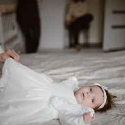 Niemowlę w białej sukience z koronką leżące na wzorzystej pościeli, przygotowywane do sesji zdjęciowej, widoczne ręce osoby poprawiającej sukienkę, w tle łóżeczko i inne osoby.