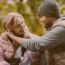 Uśmiechnięta blondynka w różowej kurtce i mężczyzna w czapce patrzą na siebie z czułością na tle jesiennego parku.