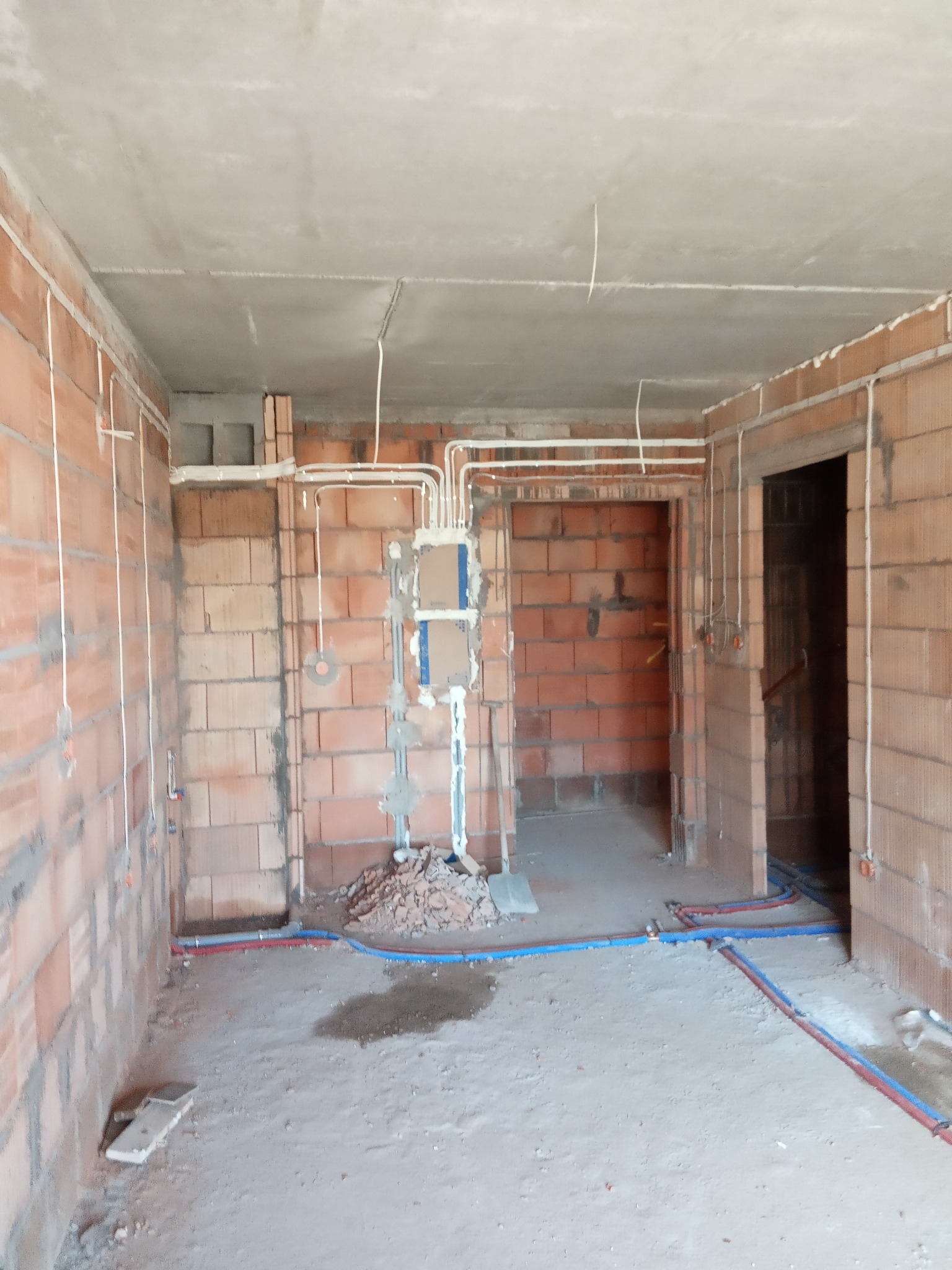 Surowy stan wnętrza z widocznymi instalacjami elektrycznymi w białych rurkach i instalacjami wodnymi w niebiesko-czerwonych rurkach, ściany z czerwonej cegły, betonowy sufit