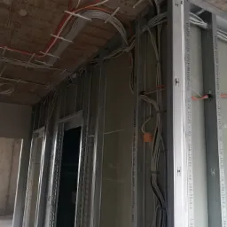 Instalacja elektryczna w budynku w trakcie budowy: widoczne przewody elektryczne prowadzone po suficie i ścianach, zamocowane do metalowej konstrukcji szkieletowej ścian działowych z płyt...