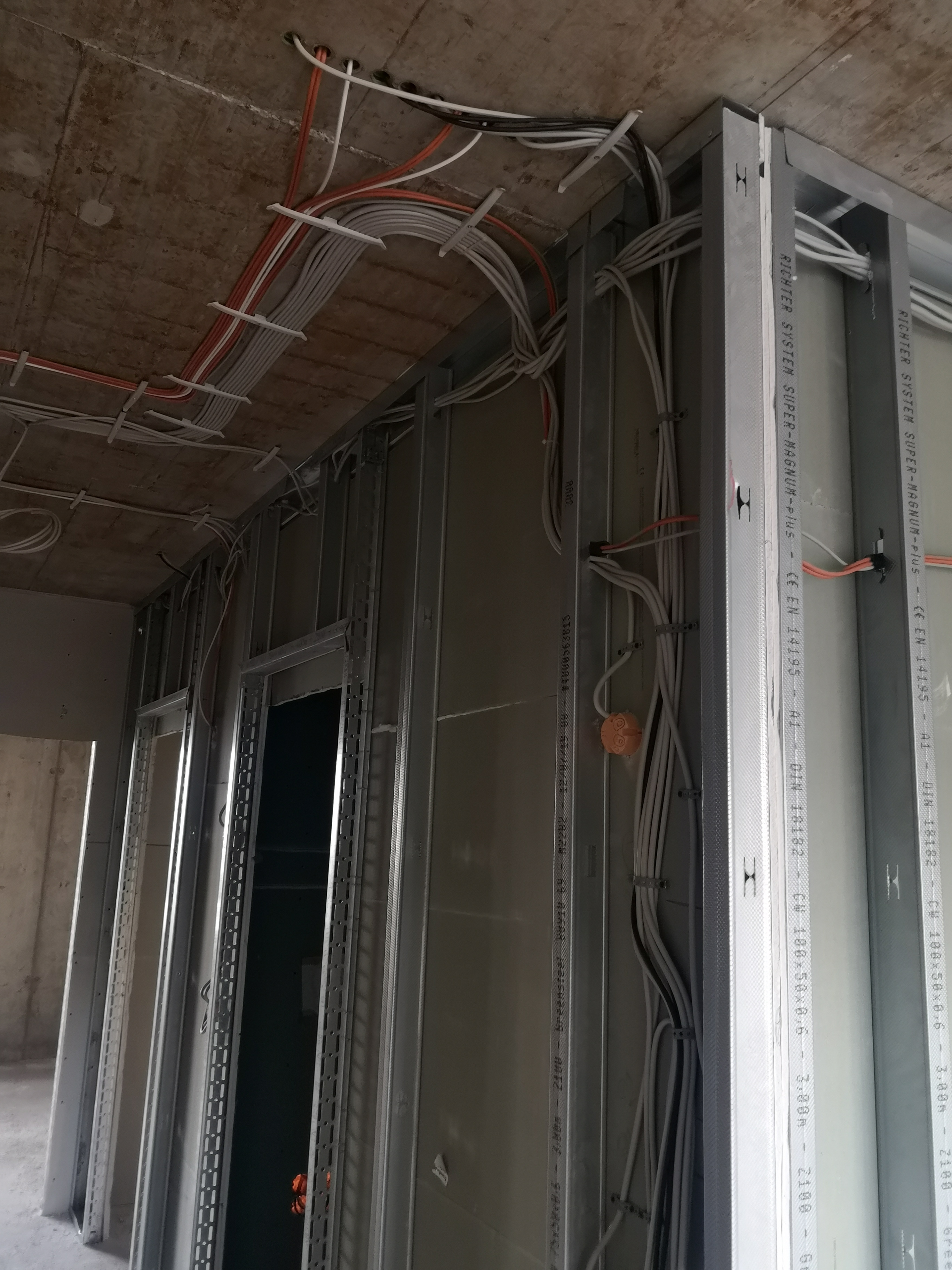 Instalacja elektryczna w budynku w trakcie budowy: widoczne przewody elektryczne prowadzone po suficie i ścianach, zamocowane do metalowej konstrukcji szkieletowej ścian działowych z płyt...