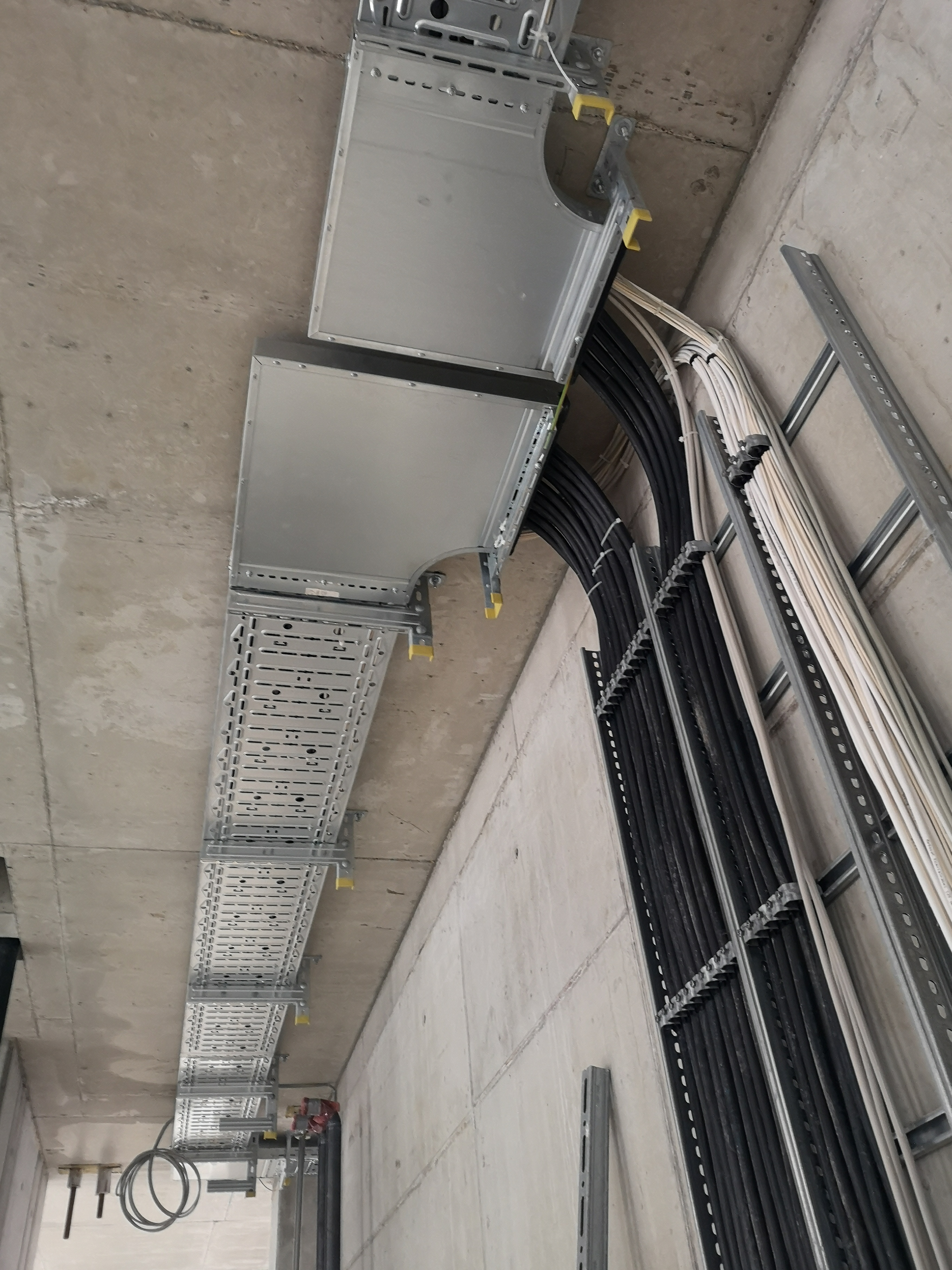 Metalowe koryta kablowe z ułożonymi przewodami elektrycznymi, zamocowane pod betonowym sufitem, widoczne z perspektywy pod kątem.