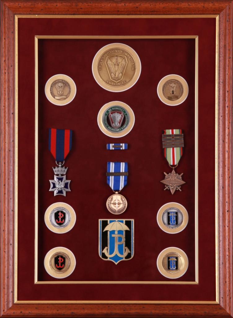 Kolekcja odznaczeń wojskowych i medali w ramce z bordowym tłem, prezentująca pamiątki związane z jednostką wojskową, w tym odznaki, medale i baretki.
