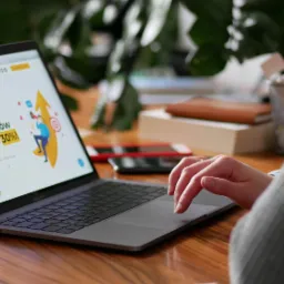 Kobieta korzysta z laptopa, na ekranie widoczna strona internetowa z hasłem 'Zwiększymy liczbę twoich klientów minimum o 30%!'. Na biurku telefon i przybory biurowe.