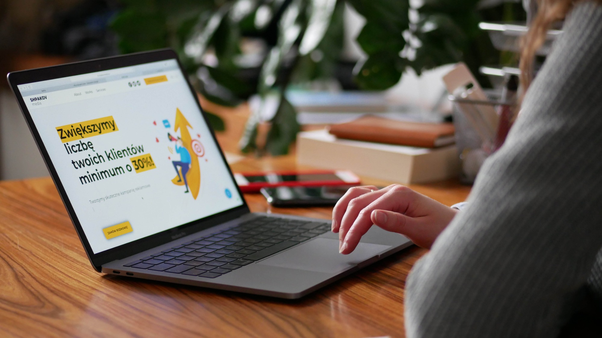 Kobieta korzysta z laptopa, na ekranie widoczna strona internetowa z hasłem 'Zwiększymy liczbę twoich klientów minimum o 30%!'. Na biurku telefon i przybory biurowe.