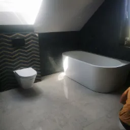 Nowoczesna łazienka na poddaszu z wolnostojącą białą wanną, podwieszaną toaletą na tle ściany w chevrony, szare płytki podłogowe i okno dachowe wpadające do wnętrza