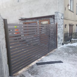 Kamil Kapała Kam-Spaw - Nowoczesna brama metalowa z poziomymi lamelami w kolorze grafitowym, zamontowana przy posesji z elewacją w odcieniach szarości, na lekko ośnieżonym podjeździe z kostki brukowej.